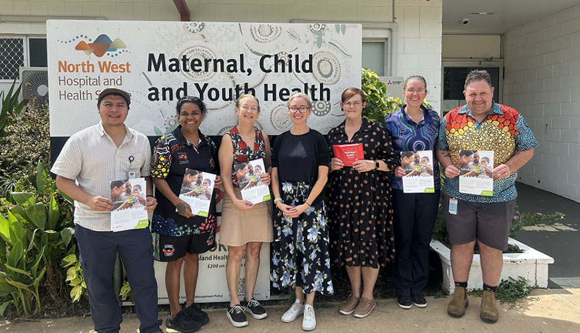 A diverse group of people stands in front of a sign reading Maternal Child and Your Health, promoting health awareness.