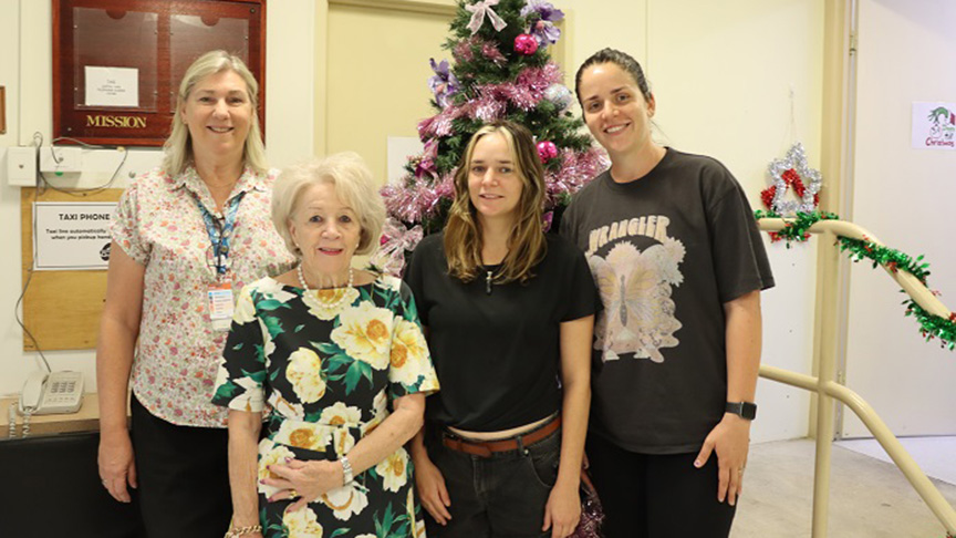 Three women and a man stand together in front of a decorated Christmas tree, smiling and celebrating the holiday season.