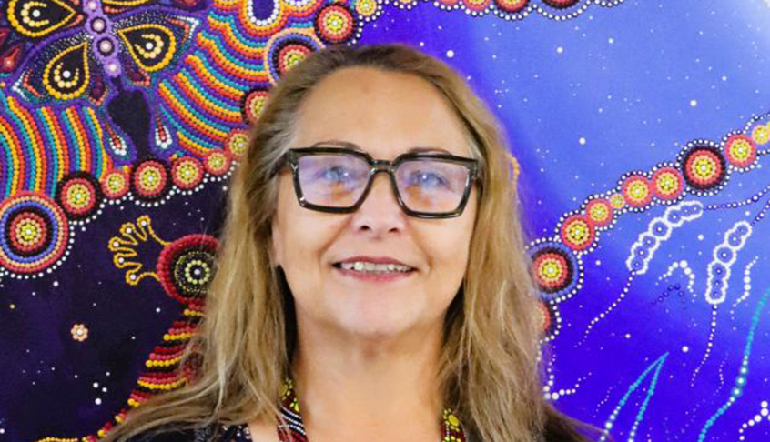 A woman wearing glasses and a necklace stands in front of a vibrant, colorful background.