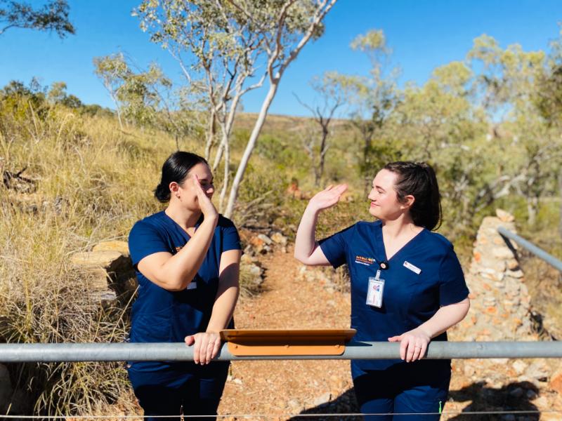 Two women in scrubs stand on a fence, smiling and engaging with each other in a friendly manner.